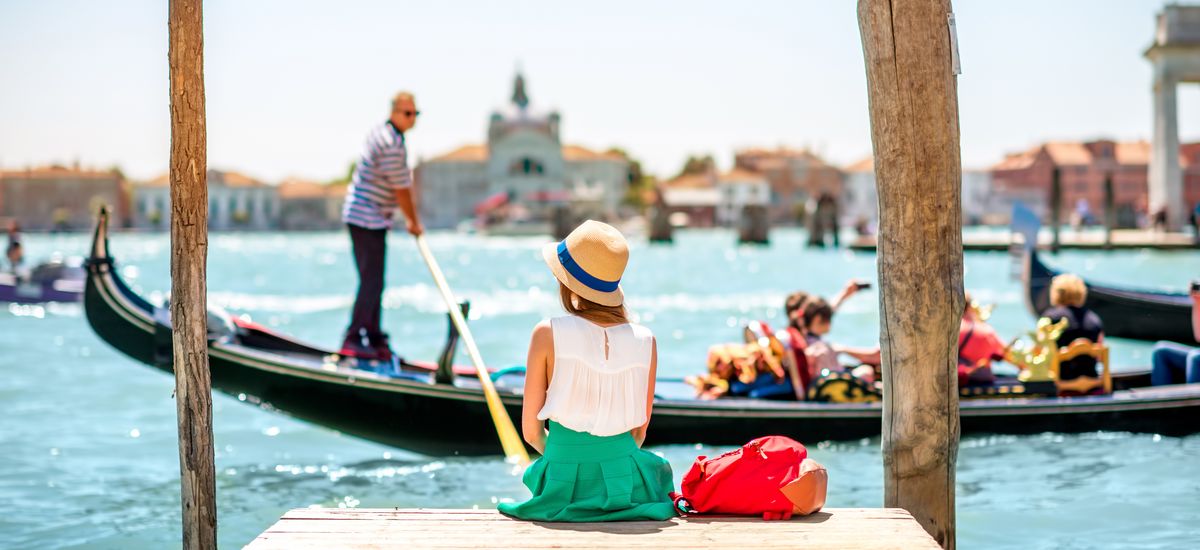 Young female traveler sitting on the pier and enjoying beautiful view on venetian chanal with gondolas floating in Venice