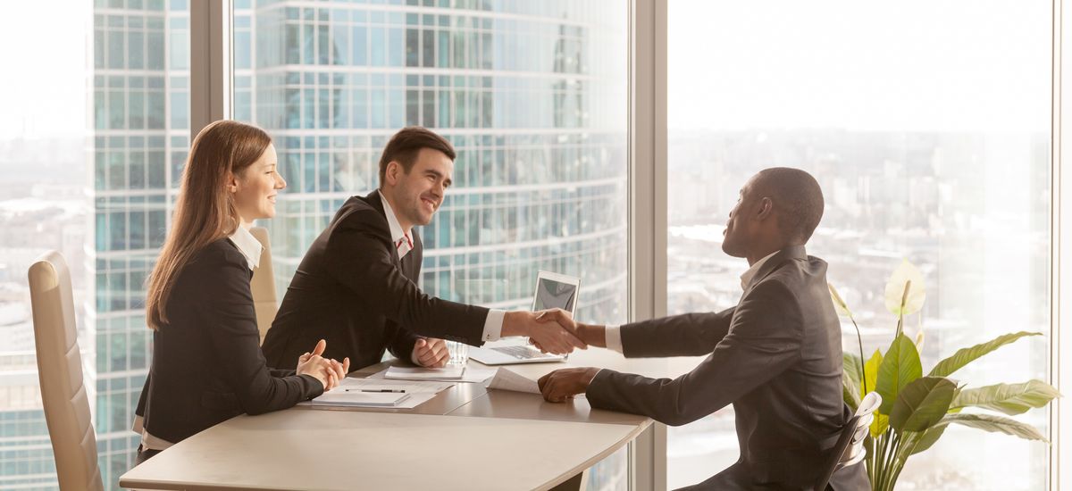 Friendly caucasian employers and confident african-american applicant handshaking during job interview sitting at office desk with big large window urban buildings cityscape at background, side view