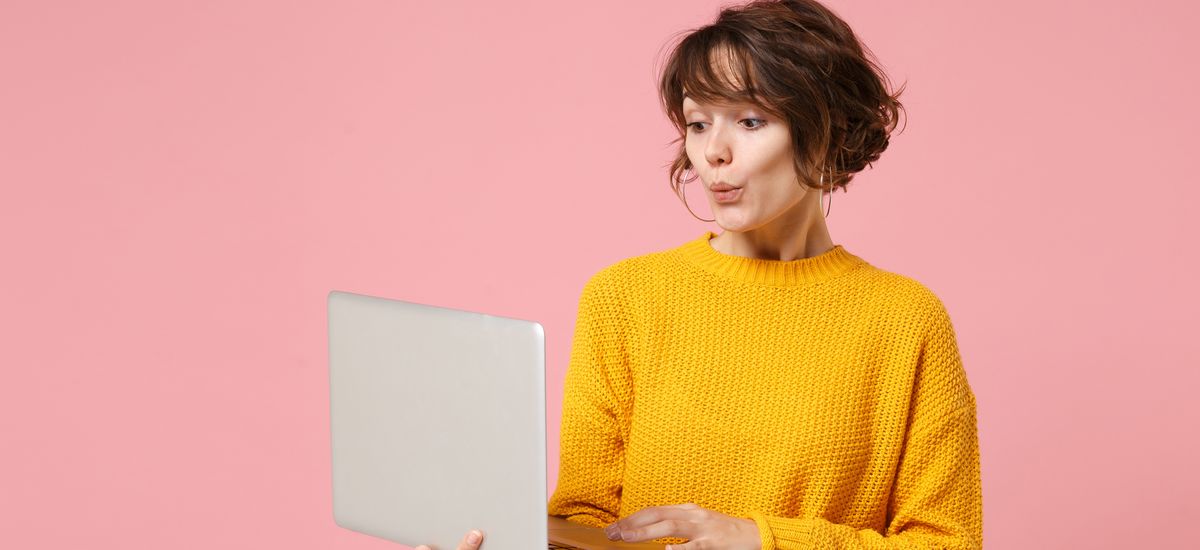 Amazed young brunette woman girl in yellow sweater posing isolated on pastel pink background studio portrait. People lifestyle concept. Mock up copy space. Holding and working on laptop pc computer.