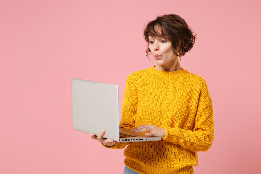 Amazed young brunette woman girl in yellow sweater posing isolated on pastel pink background studio portrait. People lifestyle concept. Mock up copy space. Holding and working on laptop pc computer.