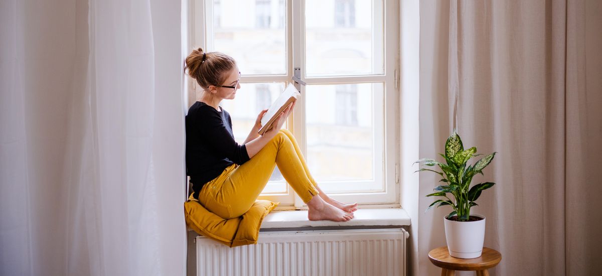 A young female student with a book sitting on window sill, studying.