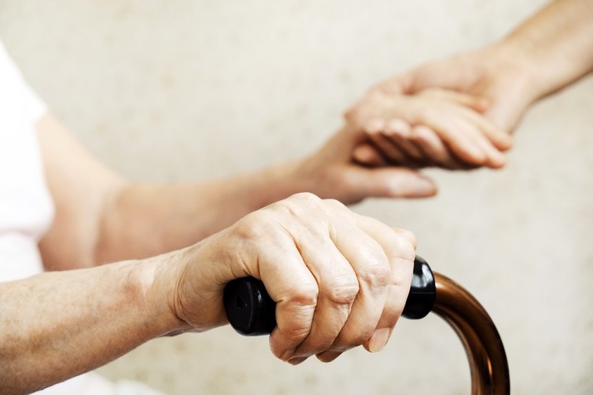Senior woman holding quad cane handle in elderly care fecility. Hospital nurse comforting mature female in nursing home, sitting with walking stick. Background, close up on hands w/ wrinkled skin.