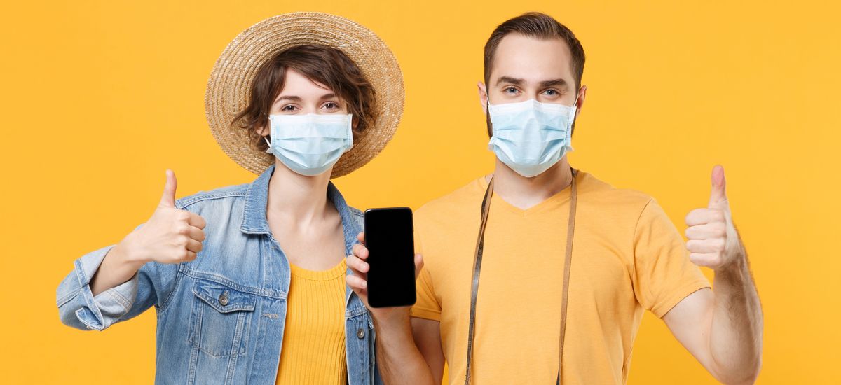 Tourists couple friends guy girl in face mask isolated on yellow background. Epidemic pandemic coronavirus 2019-ncov covid-19 flu virus concept. Hold mobile phone with blank screen showing thumbs up.