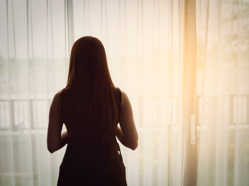 Back view of woman standing beside the windows with sunlight in 