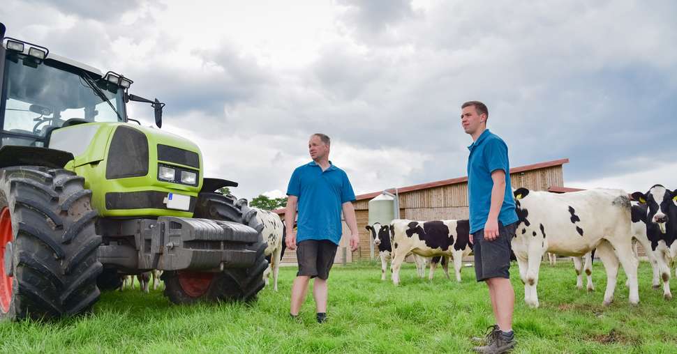 Landwirt mit Sohn und Trecker mit Rindern in der Weide, Rindviehstall im Hintergrund